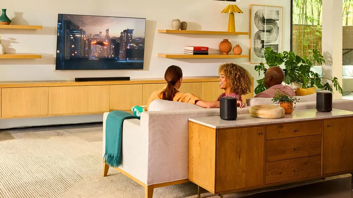 Two black JBL Cove M1 speakers on a sideboard behind a sofa, with a JBL soundbar under a wall-mounted TV